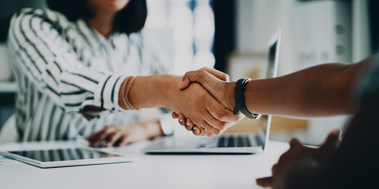 Two women shaking hands while seated at a table with a laptop.